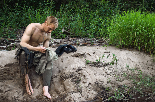 jonge man in een oude militaire uniform ontdaan aan de taille zit op een zandstrand - Foto, afbeelding