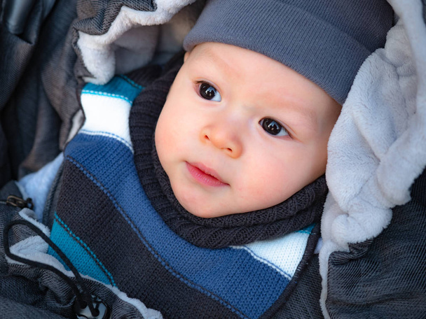 Close up portrait of sweet newborn boy wrapped in winter envelope outdoors, Happy laughing baby enjoying a walk in a snowy winter park sitting in a warm stroller with sheepskin hood. - Photo, Image