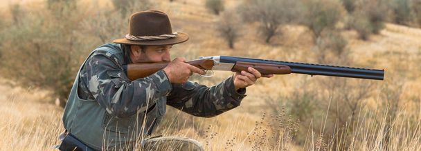 Silhueta de um caçador com uma arma. Um caçador de chapéu com uma arma em camuflagem e um colete reflexivo na estepe
 - Foto, Imagem