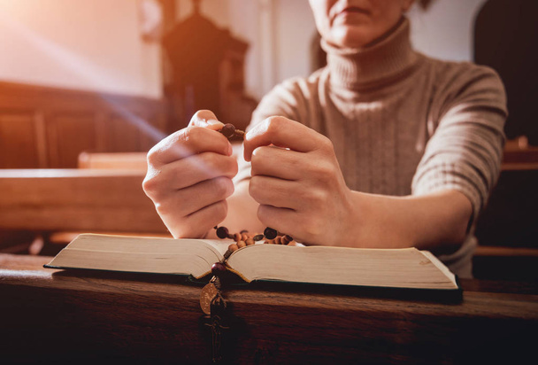 Christian woman praying in church. Hands crossed and Holy Bible on wooden desk. Background - Zdjęcie, obraz