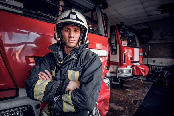 Μια αυτοπεποίθηση πυροσβέστης φορούσε προστατευτική στολή στέκεται δίπλα σε ένα πυροσβεστικό όχημα σε ένα γκαράζ του μια πυροσβεστική, σταυρωμένα χέρια και κοιτάζοντας μια φωτογραφική μηχανή - Φωτογραφία, εικόνα