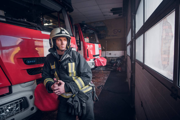 Young fireman wearing protective uniform standing next to a fire engine in a garage of a fire department - Fotoğraf, Görsel