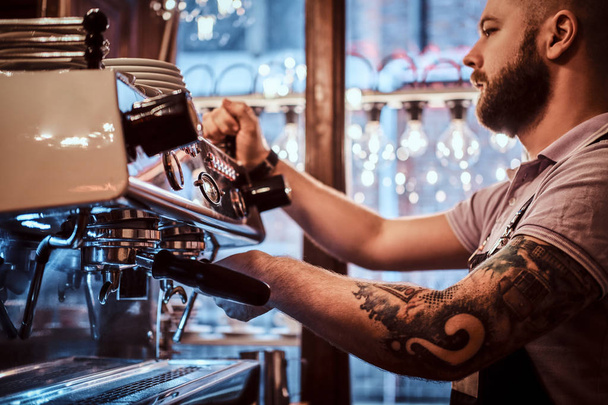 Τατουάζ barista με στιλάτα γένια και το χτένισμα που εργάζεται πάνω σε μια μηχανή του καφέ σε μια καφετέρια ή εστιατόριο - Φωτογραφία, εικόνα