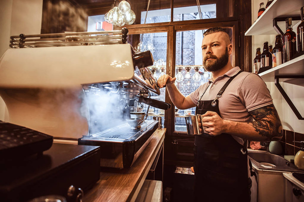 Tetovaný barista s stylové vousy a účes pracuje na automatu na kávu v kavárně nebo restauraci - Fotografie, Obrázek