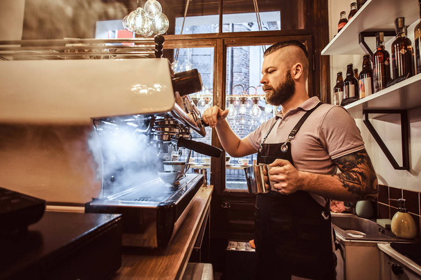 Τατουάζ barista με στιλάτα γένια και το χτένισμα που εργάζεται πάνω σε μια μηχανή του καφέ σε μια καφετέρια ή εστιατόριο - Φωτογραφία, εικόνα