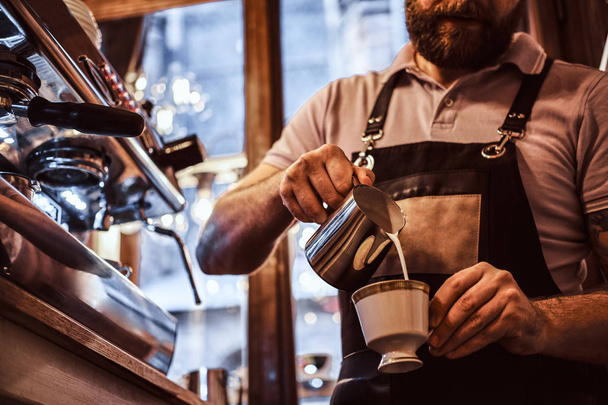 Barista vestindo avental fazendo um cappuccino, derramando leite em uma xícara em um restaurante ou café
 - Foto, Imagem