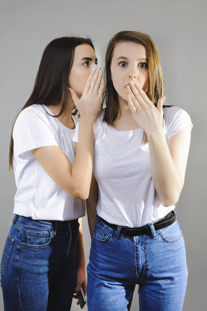 Όμορφη νεαρή ευρωπαϊκή μελαχρινή κουτσομπολεύουν στην φίλη της τρώνε ενώ που ψάχνει κατάπληκτος σε κάμερα ντυμένος στα λευκά πουκάμισα και τζιν ενάντια σε έναν τοίχο λευκό. - Φωτογραφία, εικόνα