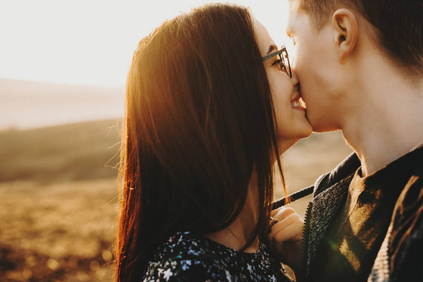 Mooie jonge vrouw die het dragen van bril glimlachen terwijl het proberen om het kussen van haar vriendje, terwijl hij weg tegen zonsondergang kijkt. - Foto, afbeelding