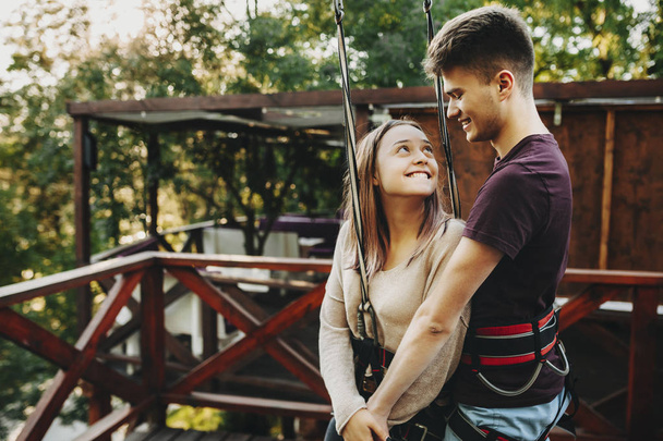 Amazing young couple looking one to another smiling with love while being equipped for rope slide while traveling together. - Photo, Image