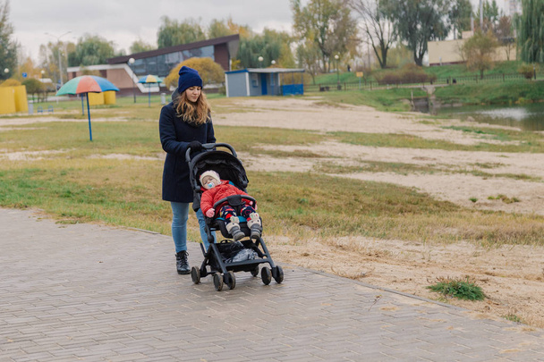 Una mujer camina en el parque con un cochecito y un niño pequeño cerca del lago
. - Foto, Imagen