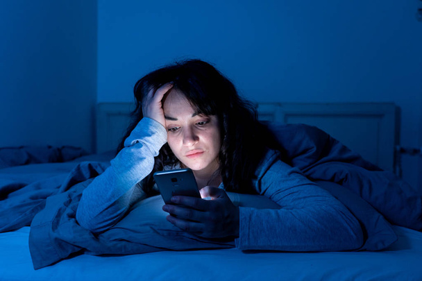 Attractive latin woman addicted to mobile phone and internet late at nigh in bed looking sleepless - Zdjęcie, obraz