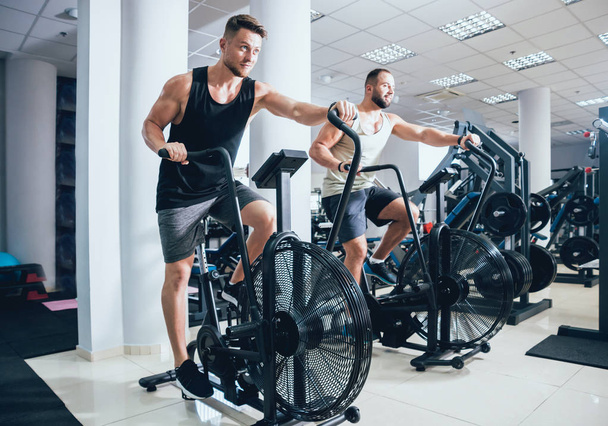 Young men with muscular body using air bike for cardio workout at cross training gym. Fitness concept. - Fotoğraf, Görsel