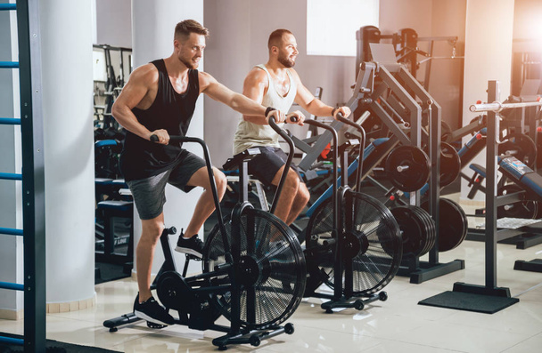 Young men with muscular body using air bike for cardio workout at cross training gym. Fitness concept. - Fotografie, Obrázek