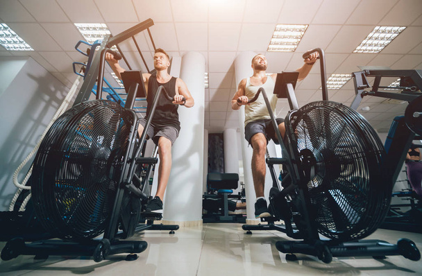 Young men with muscular body using air bike for cardio workout at cross training gym. Fitness concept. - Fotoğraf, Görsel
