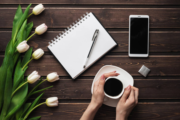 White tulips on a wooden table with an empty notebook and smartphone and a Cup of coffee in womens hands - Fotoğraf, Görsel