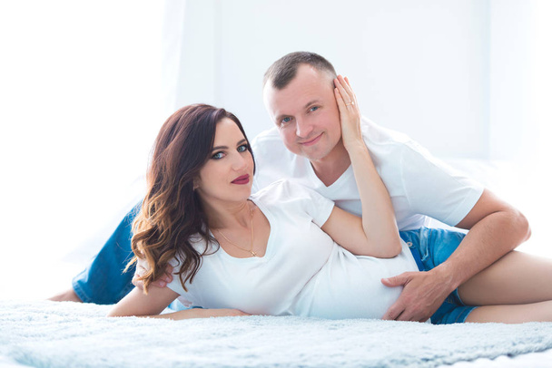 Cute pregnant couple woman in jeans overalls and a man lying on the bed near the window in a bright room - Photo, image