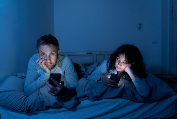 Life style portrait of young bored couple in bed at night on smart phones obsessed with games, social media, apps ignoring each other. Relationship communication problems and phone addiction concept. - Foto, afbeelding