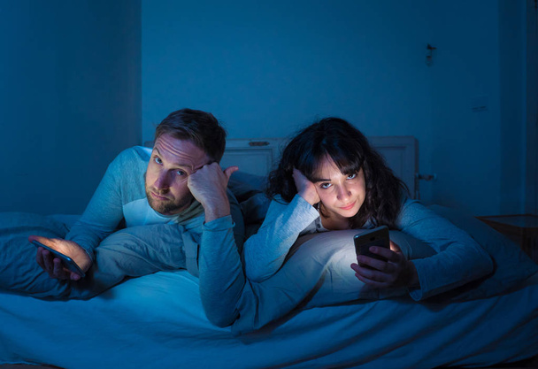 Life style portrait of young bored couple in bed at night on smart phones obsessed with games, social media, apps ignoring each other. Relationship communication problems and phone addiction concept. - Фото, зображення