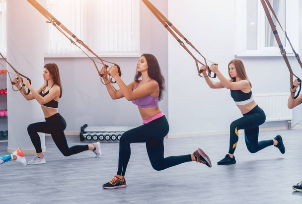 Beautiful young sports women are working out with TRX in gym. Light background - Foto, immagini