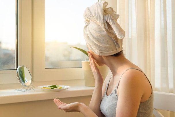 Young woman at home near the window looking in mirror, caring for face with cream and making mask with cucumber and lemon, towel on head. Skin care, cosmetics, cosmetology, dermatology. - Фото, зображення