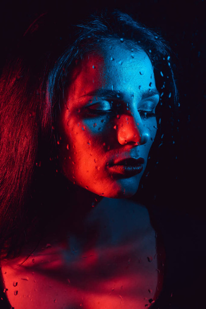 Portrait of sad young girl with red blue lighting behind glass with raindrops - Φωτογραφία, εικόνα