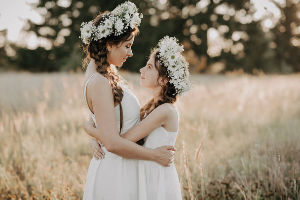 hermanas felices en vestidos blancos con coronas florales y trenzas de estilo boho en verano en un campo
 - Foto, imagen