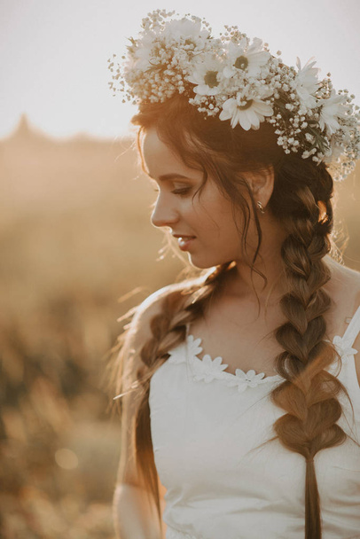 portrait of attractive girl with flower wreath and braids in white dress in summer field at sunset - Fotoğraf, Görsel