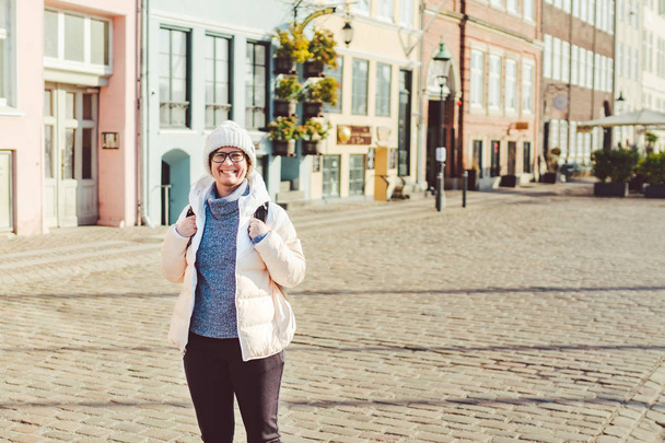 Portrait of a young Caucasian European woman tourist in glasses for the view of a white hat and a down jacket with a black backpack posing on an old Europe street in the city of Copenhagen in Denmark - Photo, Image