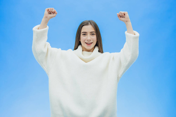 Attractive young girl. With Black Hair Celebrating Victory. Waving with his fists and a brilliant smile. On a blue background, a studio with a copy of the space. - Фото, изображение