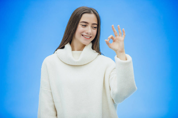 The image of a happy young lady standing on a blue background. Look into the camera. During this time she is dressed in a white warm sweater and shows various gestures. - Photo, Image