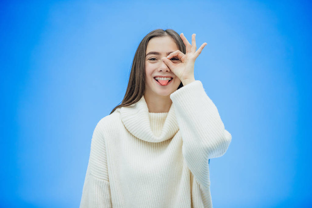 The image of a happy young lady standing on a blue background. Look into the camera. During this time she is dressed in a white warm sweater and shows various gestures. - Valokuva, kuva