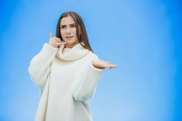 Beautiful young girl on a blue background. The girl shows a phone call with a gesture. Dressed in a white warm sweater. Has long black hair. - Фото, зображення