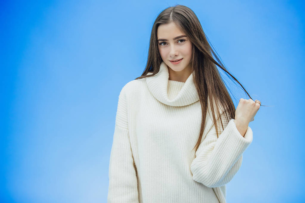 Young beautiful brunette woman on an isolated background. Hands holding hair strands. Smiling confidently and happily. On a blue background. Dressed in a white sweater. - Fotó, kép