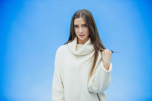 Young beautiful brunette woman on an isolated background. Hands holding hair strands. Smiling confidently and happily. On a blue background. Dressed in a white sweater. - Foto, afbeelding