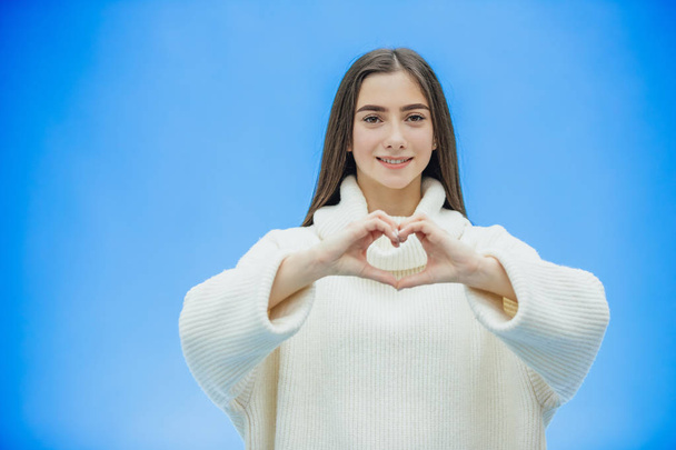 Portrait of a happy young woman wearing a white warm sweater. During this time, a hand gesture of heart shows and feels love. Isolated on a blue background. Happy and smiling. - Valokuva, kuva