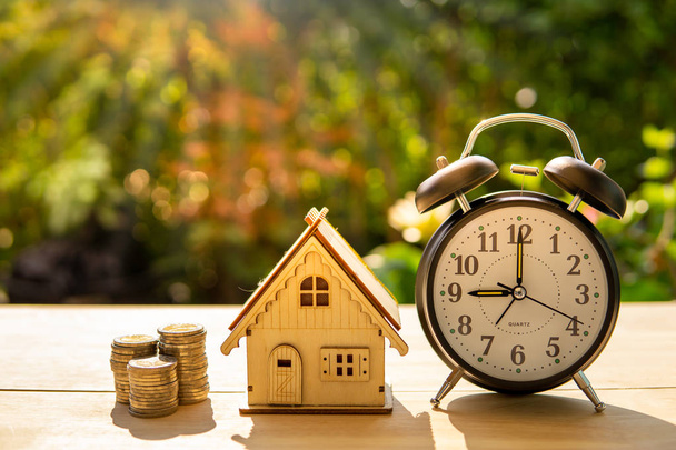 The money coins house and alarm clock stack on wood table and sunset background in the public park show savings money for house, investment, retirement and for the future and time. Business concept. - Photo, Image