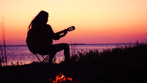 Young girl playing guitar on the beach with campfire at sunset - Кадры, видео