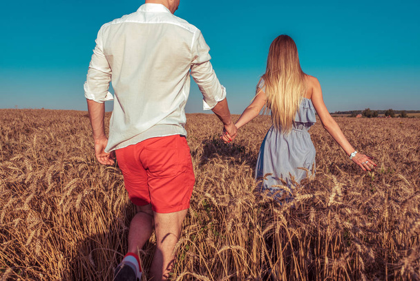 Girl in dress holds hand of man of her boyfriend husband, leads him into wheat field behind him. The guy in the shirt and red shorts. Concept follow me, walk in field in fresh air. - 写真・画像
