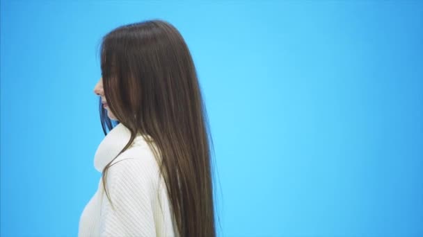 Young energetic girl stands on a blue background. During this smiles, having adjusted hair. - Filmati, video