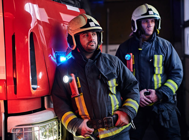 Deux pompiers portant un uniforme de protection debout à côté d'un camion de pompiers dans un garage d'un service d'incendie
. - Photo, image