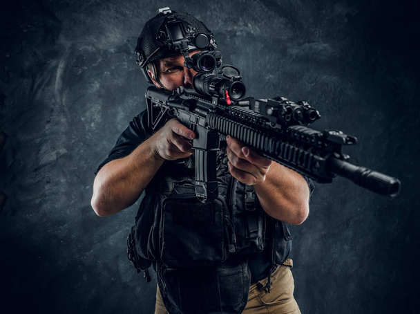 Soldado de las fuerzas especiales con chaleco antibalas y casco con visión nocturna sosteniendo un rifle de asalto. Foto del estudio contra una pared de textura oscura
 - Foto, Imagen
