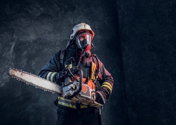 Πορτραίτο του πυροσβέστη με κράνος ασφαλείας και μάσκα οξυγόνου κρατώντας ένα αλυσοπρίονο. Στούντιο φωτογραφία σε ένα σκοτεινό υφή τοίχο - Φωτογραφία, εικόνα