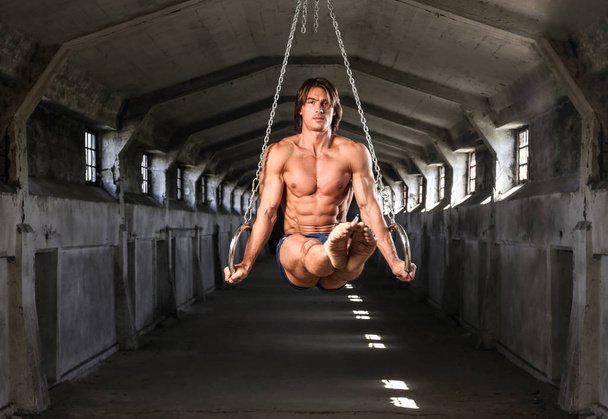 A professional sportsman with a beautiful muscular body trains on gymnastic rings in abandoned industrial building - Photo, Image