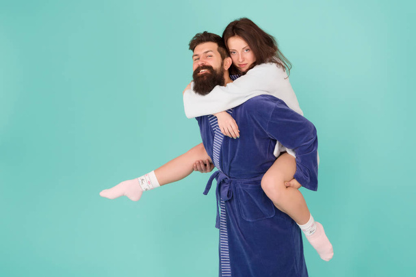Couple in bathrobes having fun turquoise background. Lets stay at home and have fun. They always have fun together. Close relationship. Handsome young man giving his girlfriend piggyback ride - Foto, immagini