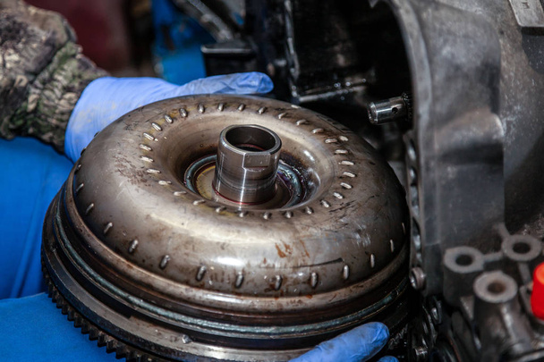 Close-up of a young male repairman in a working uniform repairing a torque converter and installing it in an automatic transmission of a supported car in an auto repair shop. The concept of car repair and car repair shop - Photo, Image