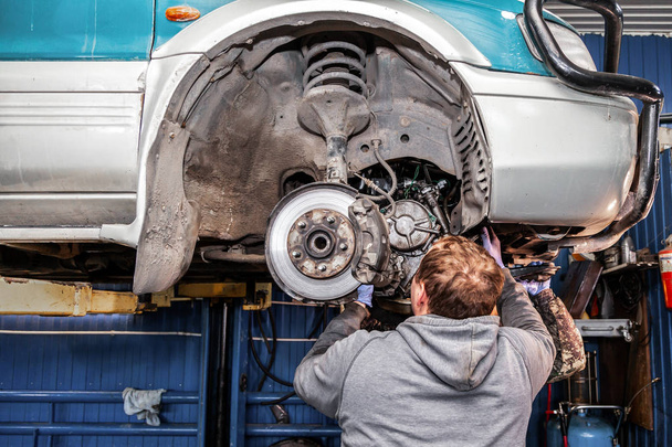 Een close-up van de reparateur van een jonge man in een uniform van de werken van auto's is het repareren van een automatische versnellingsbak en wordt geïnstalleerd in de gebruikte auto voor in een auto reparatiewerkplaats. Het concept van auto reparatie en auto reparatiewerkplaats - Foto, afbeelding