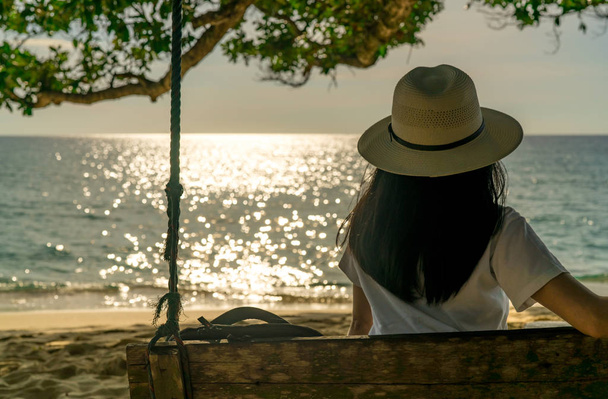 Молоді азіатські жінки сидіти і розслабитися на гойдалки на березі моря на літні канікули. Літній vibes. Жінка пам'ятки одні на відпочинок. Backpacker насолоджуватися життям в тропічний рай beach. Задній вид жінка дивитися захід сонця  - Фото, зображення