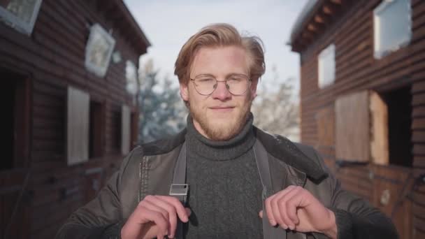 Portrait of a handsome bearded guy with glasses dressed in a warm sweater and wide-open jacket. Guy smiles, pulls up suspenders and looks around. Slow motion. - Filmagem, Vídeo