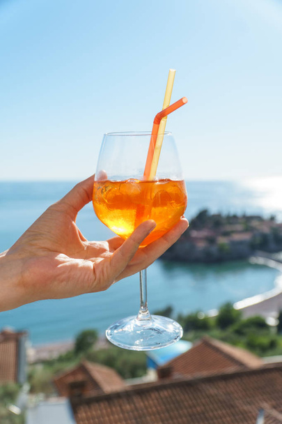ein mann hält einen orangefarbenen cocktail in einem glas mit eis und röhrchen vor dem hintergrund europäischer häuser mit roten fliesen, dem adriatischen meer und der insel sveti stefan. - Foto, Bild