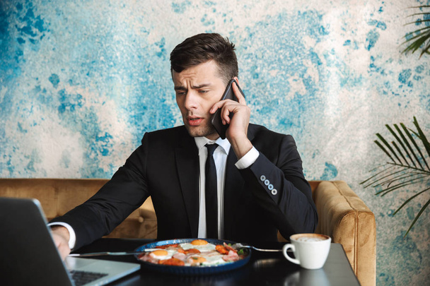 Homme d'affaires assis dans un café à l'aide d'un ordinateur portable ont un petit déjeuner ou dîner manger parler par téléphone
. - Photo, image